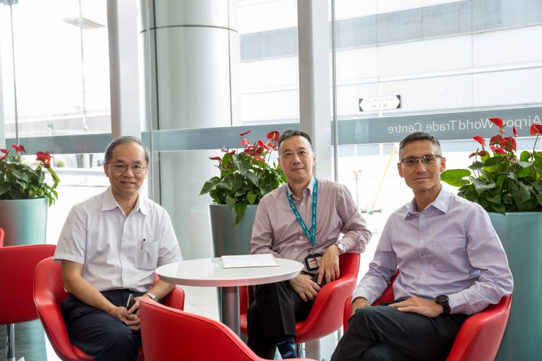 Three men seated in red chairs around a white table in a modern lobby with potted plants.