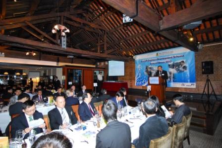 Conference attendees seated at tables listening to a presentation on a stage.