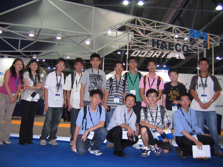 "Group of attendees posing at an aerospace conference booth with ""HAECO"" branding visible."