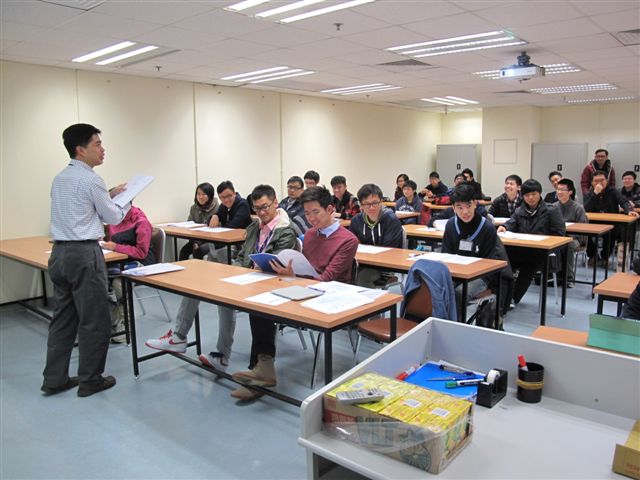 Teacher presenting to students in a classroom setting with desks and chairs.