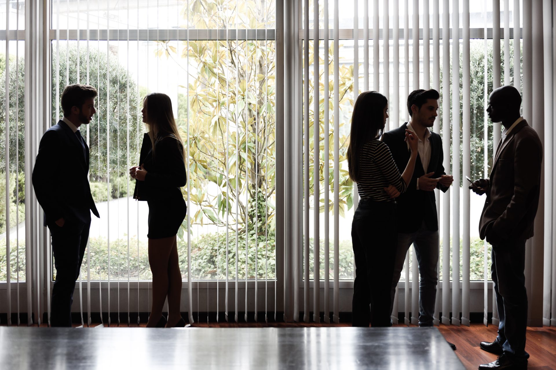 Businesspeople conversing in office near large window with vertical blinds.