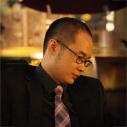 Dr. Ernest TSANG, wearing glasses and tie, looking downward in a dimly lit setting.