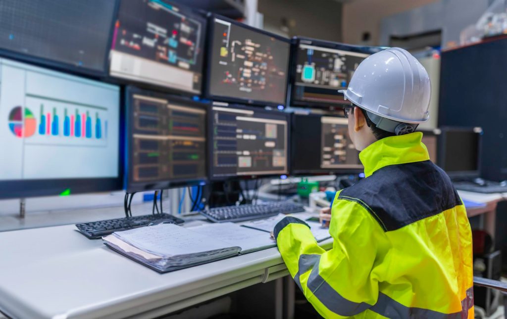 Engineer in hard hat monitoring industrial control systems on multiple screens.