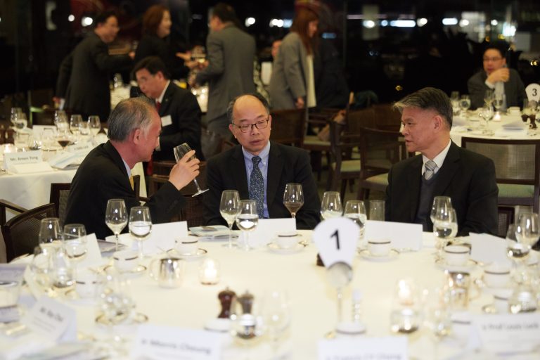 Three men in suits converse at a formal dinner table with table number one visible.