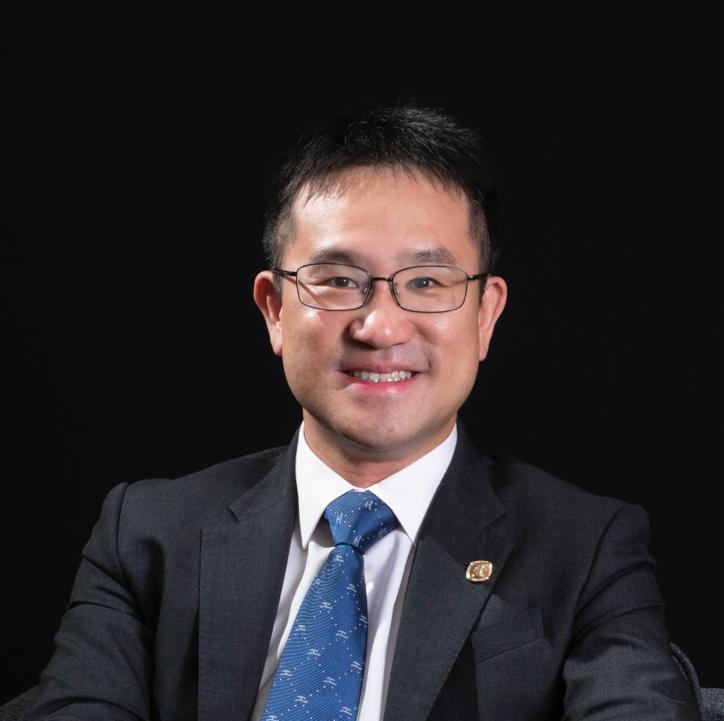 Professional headshot of a smiling man in suit and glasses against black background