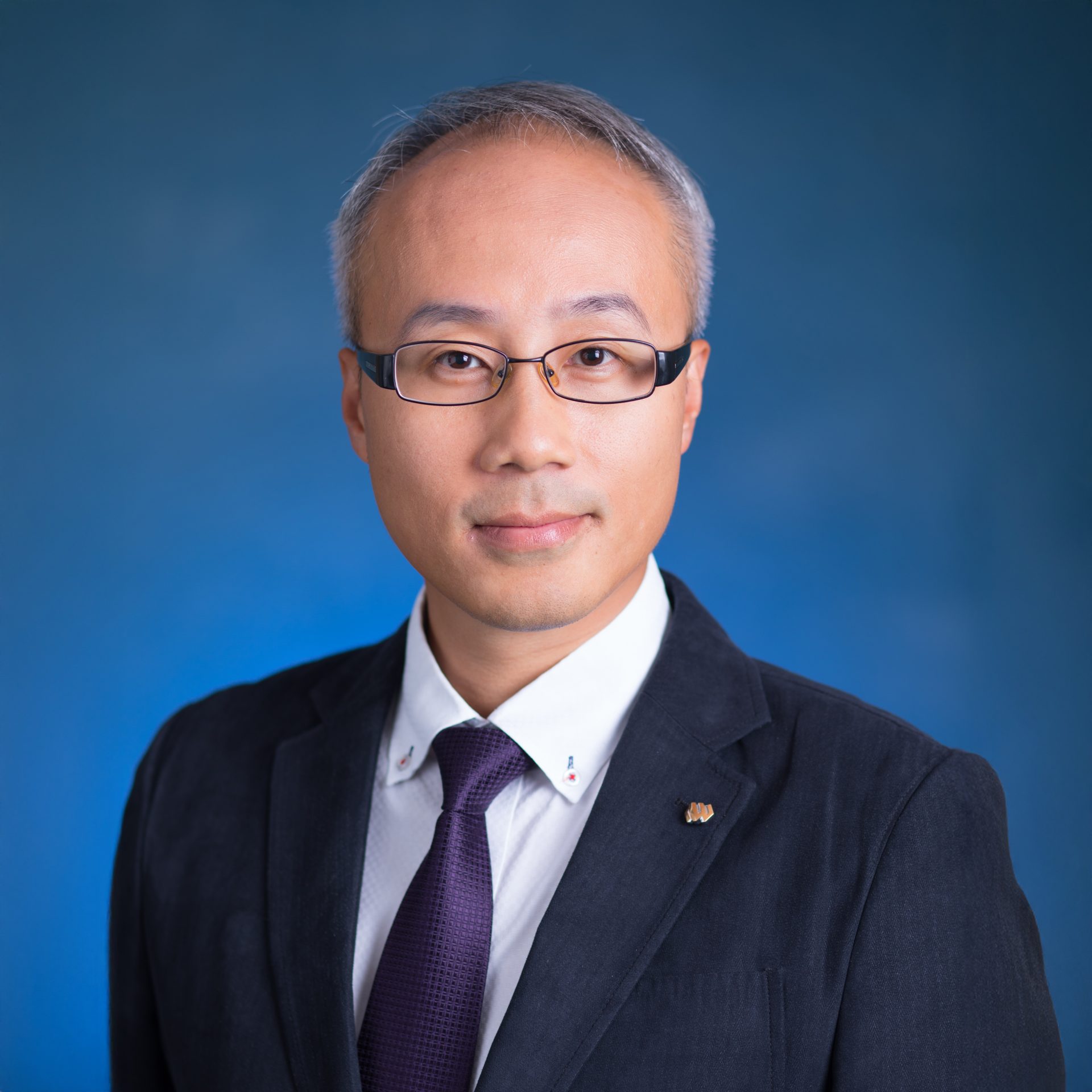 Professional headshot of a man with glasses wearing a suit and tie against a blue background.