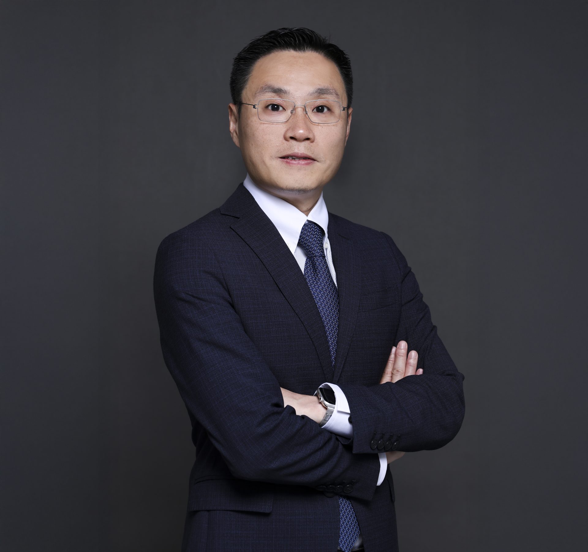Man in dark suit and glasses with arms crossed, wearing a patterned tie and smartwatch.