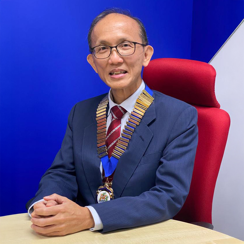 Man in suit and glasses wearing medals, seated at desk against blue background