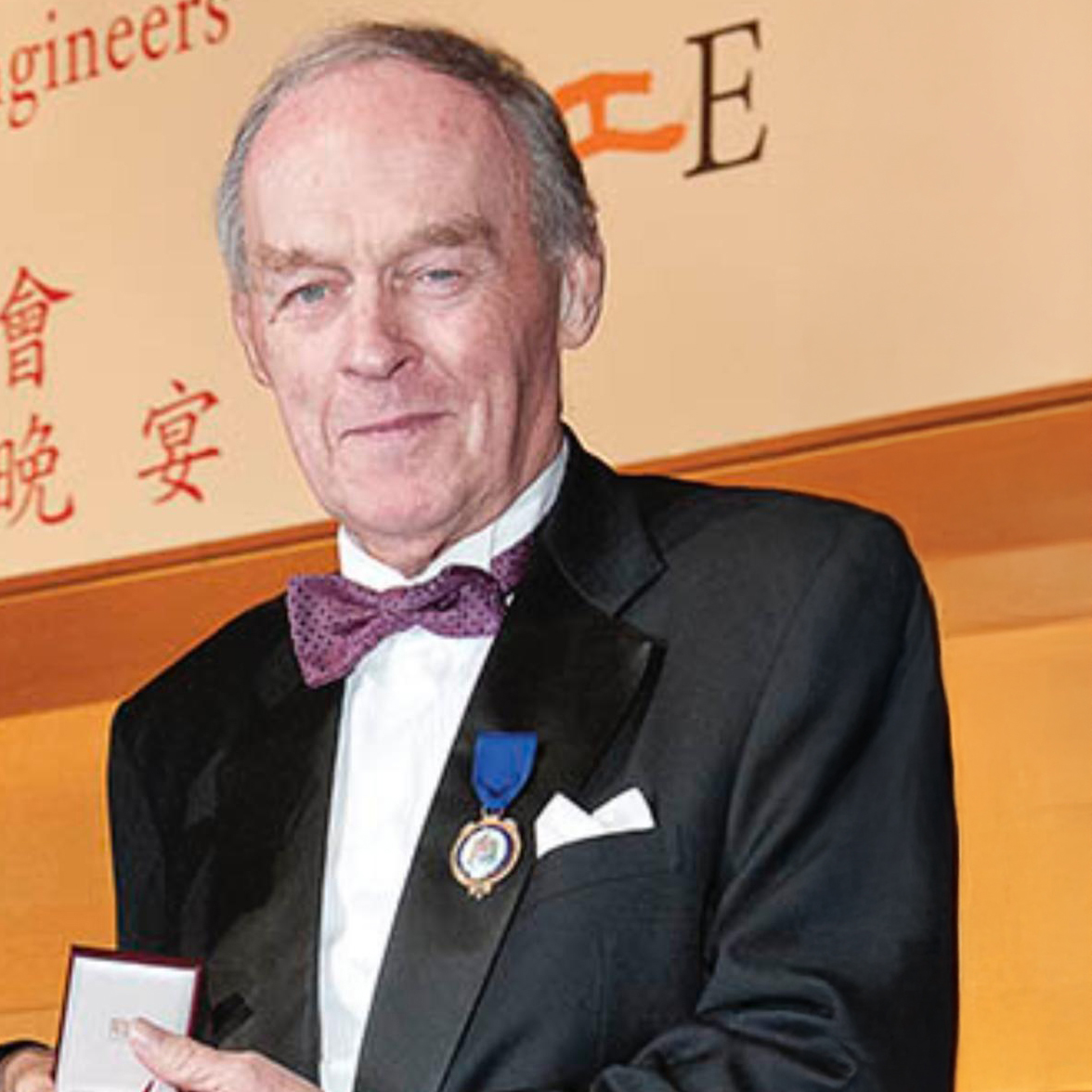 Man in tuxedo with purple bow tie and medal holding award box at event