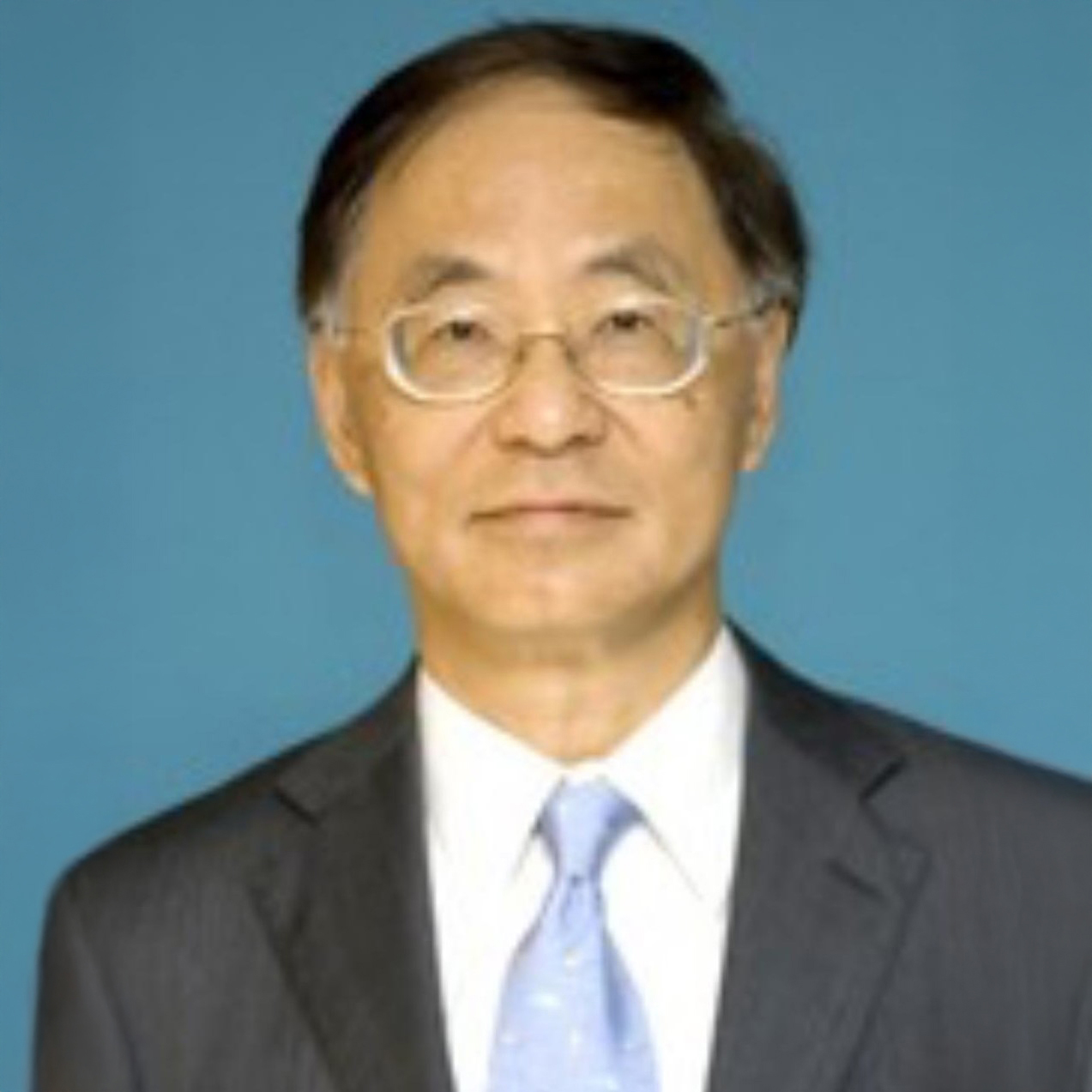 Portrait of a man with glasses wearing a suit and light blue tie against a blue background.
