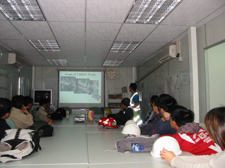 Construction workers in hard hats attend a meeting reviewing LDB201 project scope on a presentation screen.