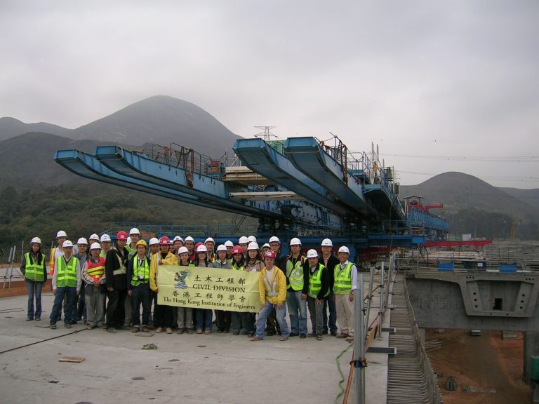 Group of engineers in safety gear posing with banner at construction site with mountains in background