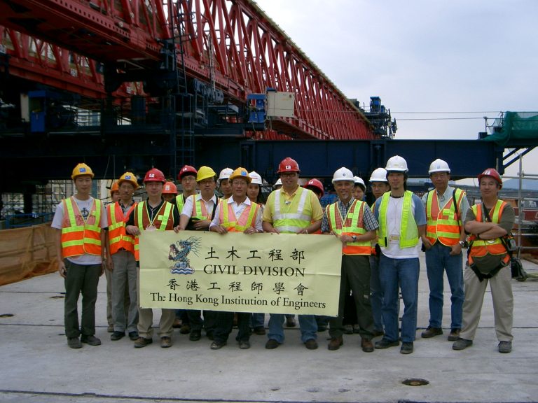 Civil engineers in safety gear pose with banner at construction site under red gantry crane.