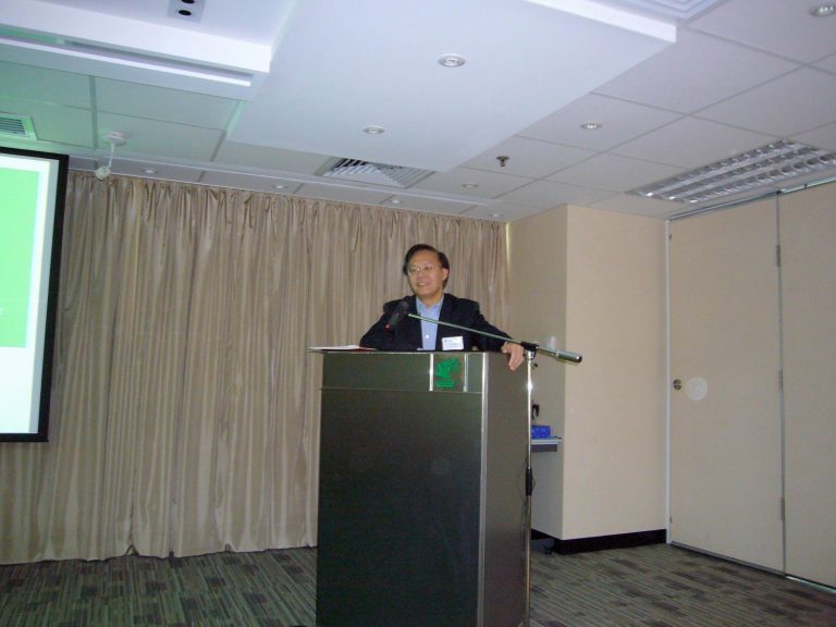 Man speaking at podium in conference room with projector screen and curtains