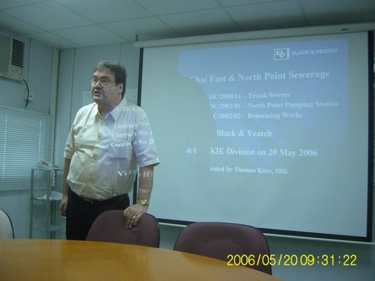 Man presenting at meeting with projector screen showing Chai East & North Point Sewerage project details.