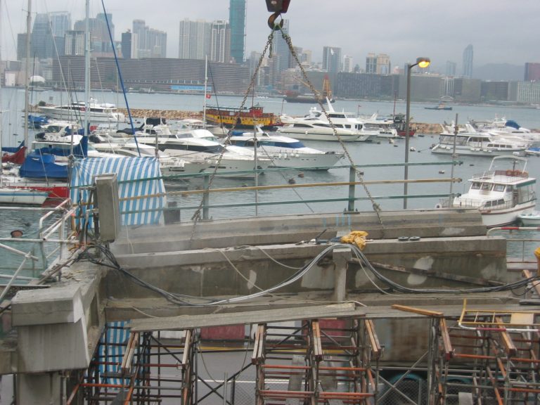 Construction crane lifting beam at harbor with boats and city skyline in background