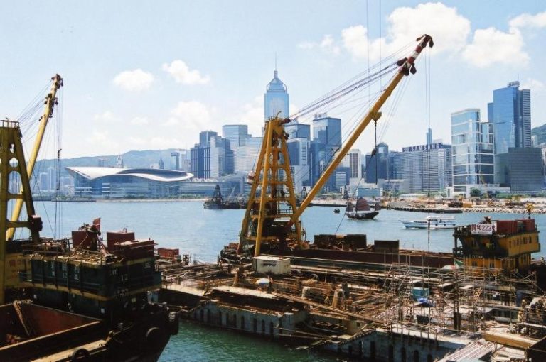 Construction cranes and barges in harbor with Hong Kong skyline in background