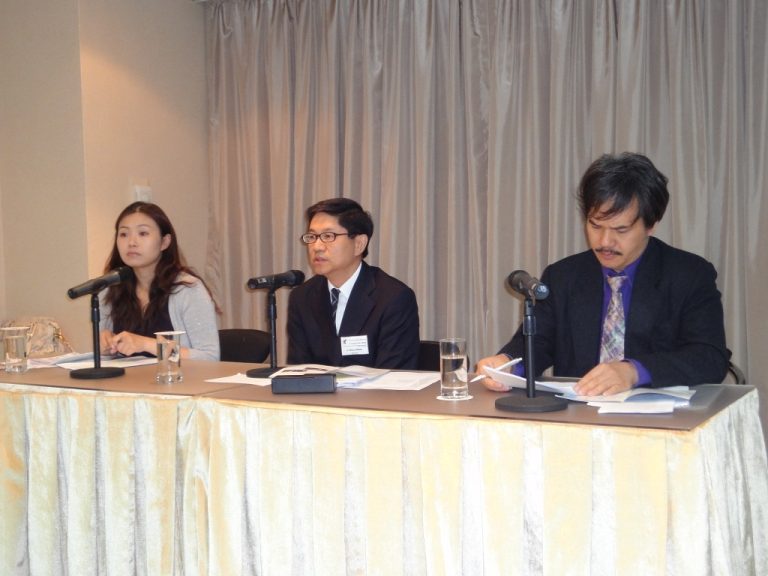 Three people seated at a table with microphones and papers, appearing to be at a press conference or panel discussion.