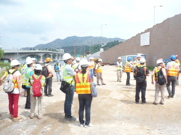 Construction workers in safety vests and hard hats gathered outdoors near a bridge and retaining wall.