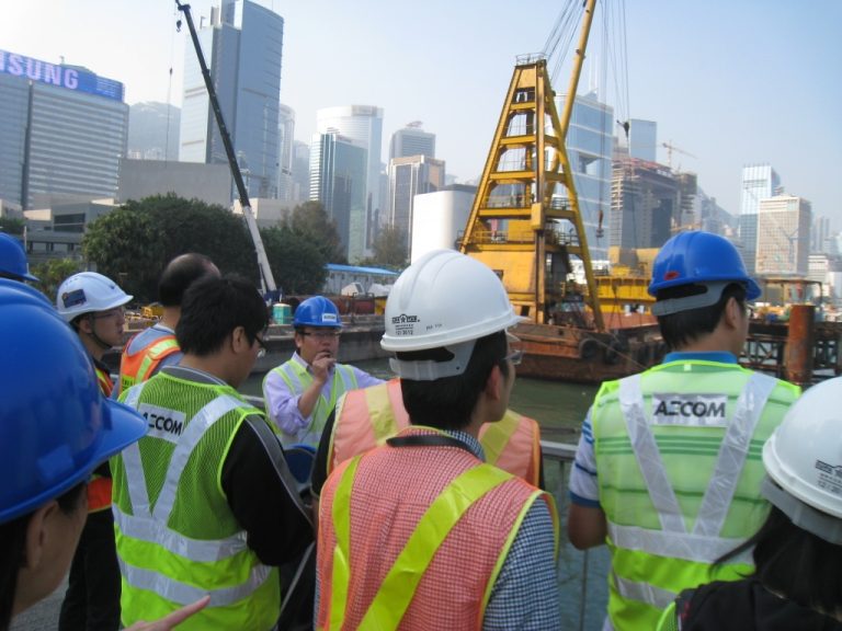 Construction workers in hard hats and safety vests listening to a site supervisor near a crane and city skyline.