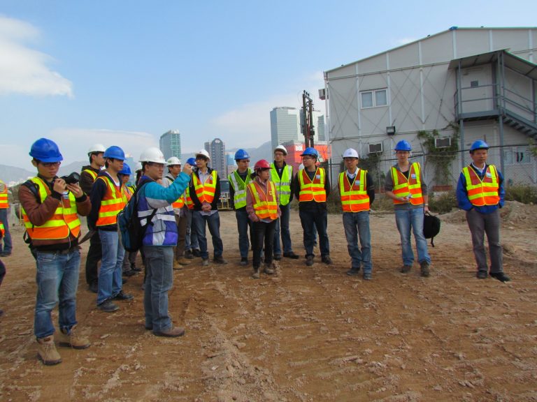 Construction workers in safety vests and hard hats gather on a dirt site with city skyline in background.