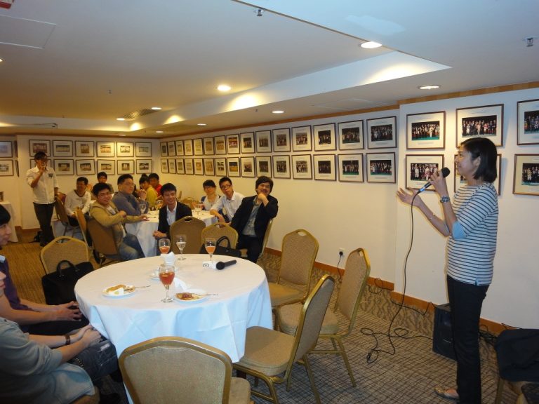 Woman speaking into microphone at a round table with seated audience in a dining room.