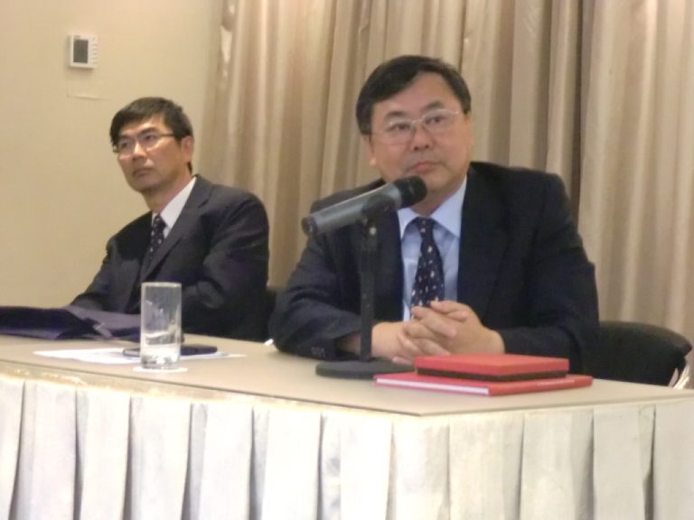 Two men in suits seated at a table with microphones and red folders.
