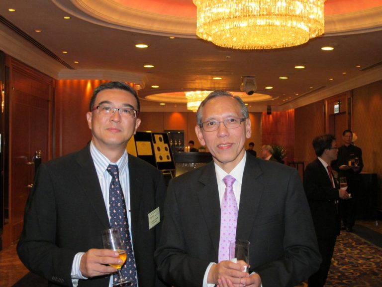 Two men in suits holding drinks at a formal event under chandeliers.