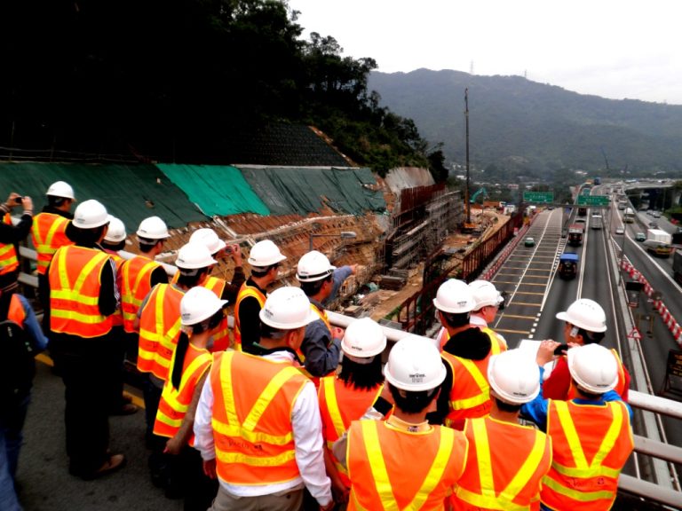 Construction workers in hard hats and safety vests observe an active highway construction site with traffic.