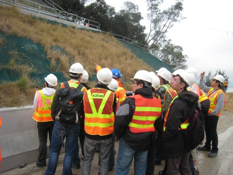 Construction workers in safety vests and hard hats inspect a landscaped embankment.