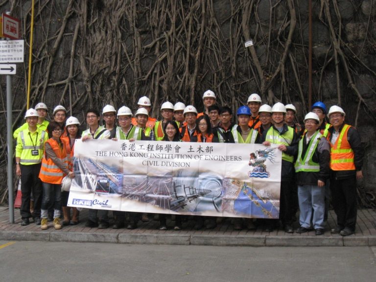 Group of civil engineers in safety gear holding HKIE Civil Division banner against tree roots.