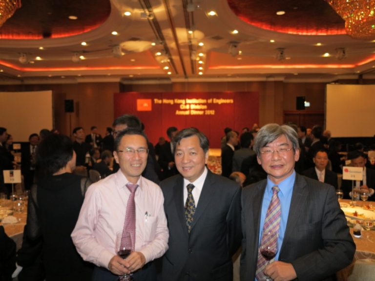 Three men smiling at an annual dinner event, holding wine glasses.