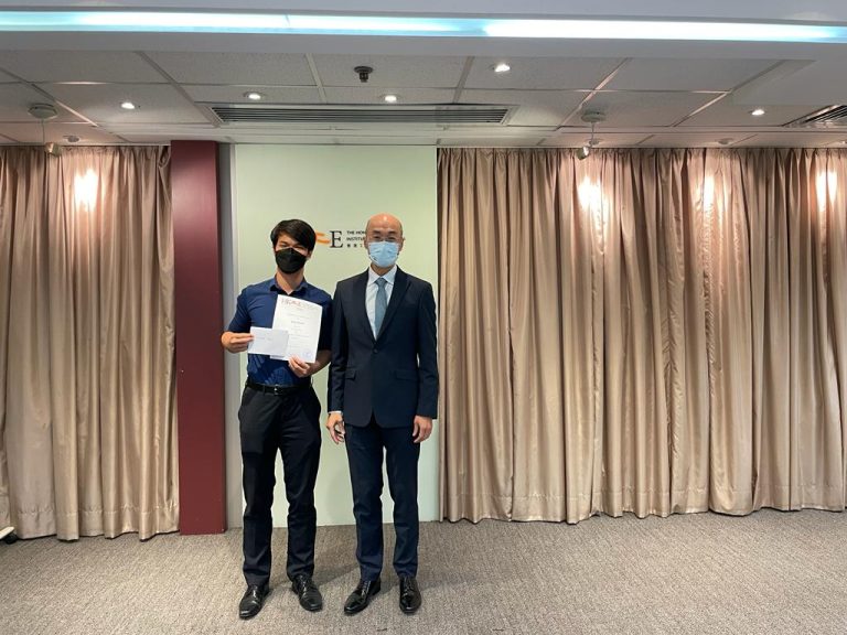 Two men in masks stand indoors, one holding a certificate, in front of curtains and a logo wall.