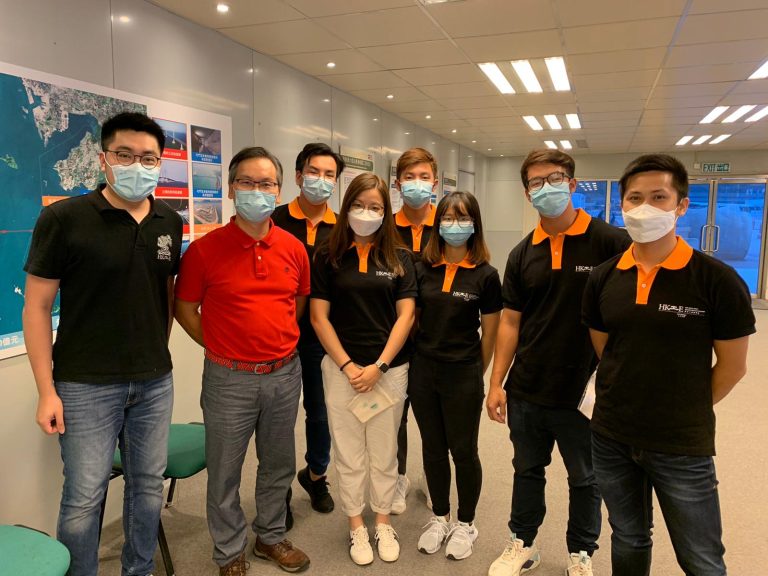 Group of eight people wearing face masks and polo shirts posing for a photo indoors.