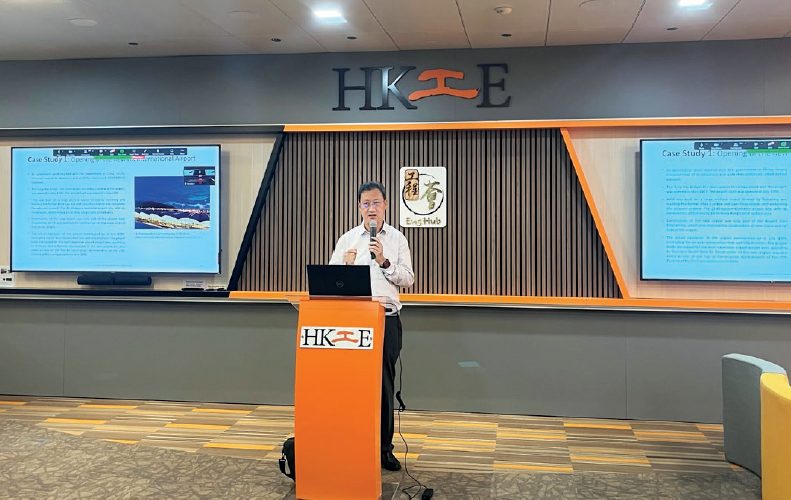 Man presenting at HKIE event with laptop and microphone, two presentation screens visible