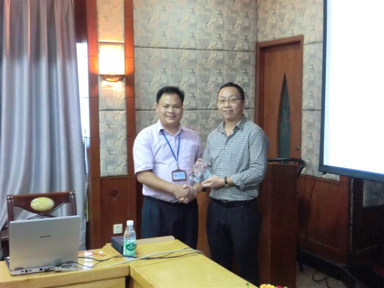 Two men shaking hands in a conference room, one holding a trophy.