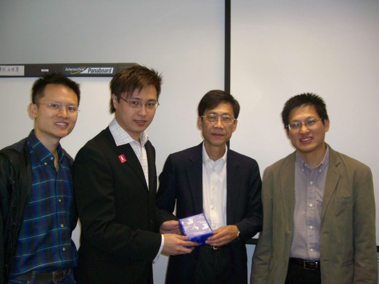 Four men posing together, two presenting a blue gift box in front of an interactive whiteboard.
