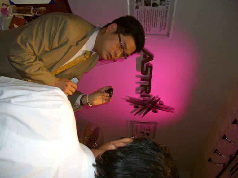 Man in suit and glasses demonstrating product at ASTRI booth with pink lighting.