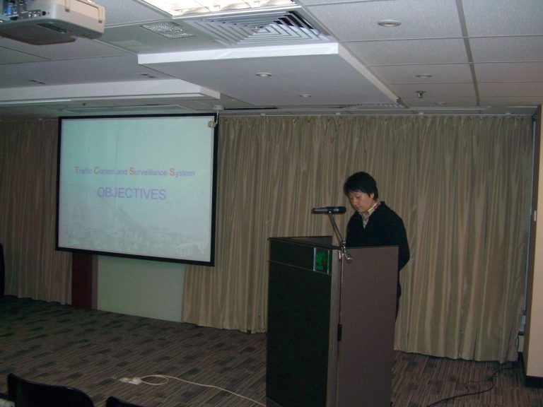 "Person presenting at podium with screen displaying ""Traffic Control and Surveillance System OBJECTIVES"