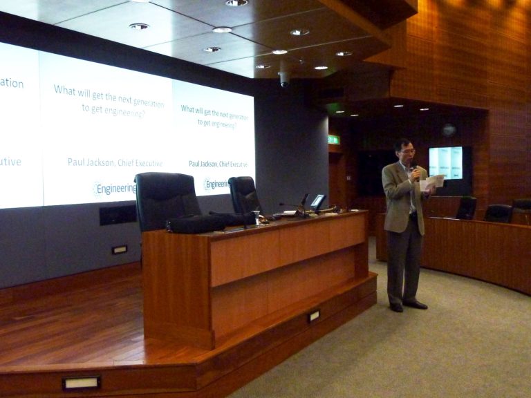  Man in a suit speaks at a wood-paneled lecture hall with a presentation slide about engineering displayed on a screen.