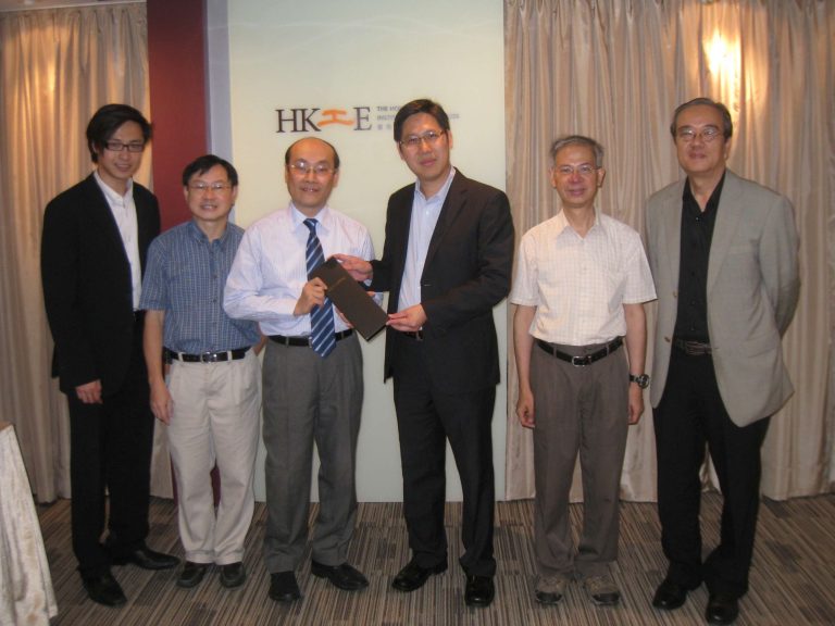 Six men in business attire pose for a photo at an event, with two exchanging a plaque.
