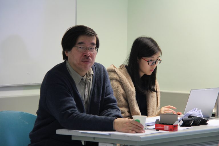 Man and woman sitting at desk, woman typing on laptop, man holding coffee cup.