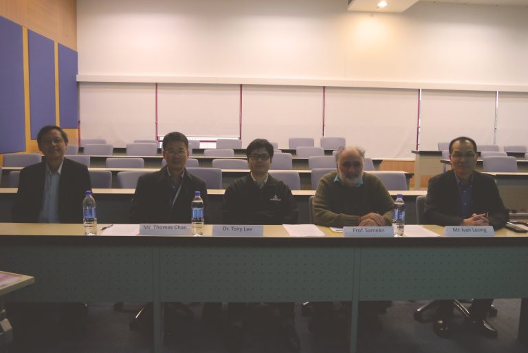 Four men seated at a conference table with nameplates in a lecture hall.