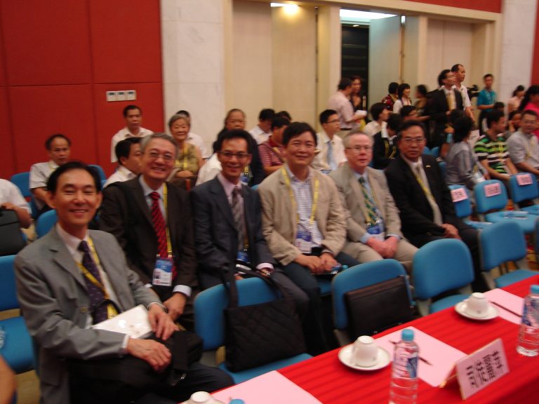 Audience at conference seated in blue chairs, smiling and wearing lanyards.