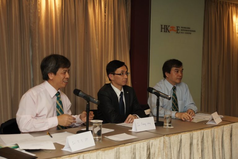 Three men in business attire seated at a conference table with microphones and name cards.