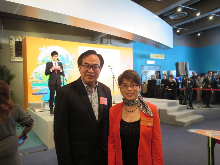 Two individuals posing for a photo at an event with a speaker on stage behind them.