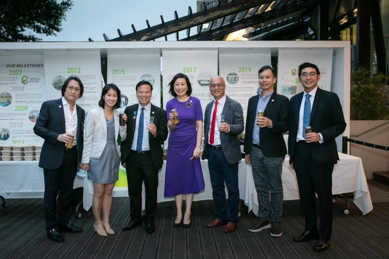 Group of seven professionals posing with drinks in front of company milestone display.