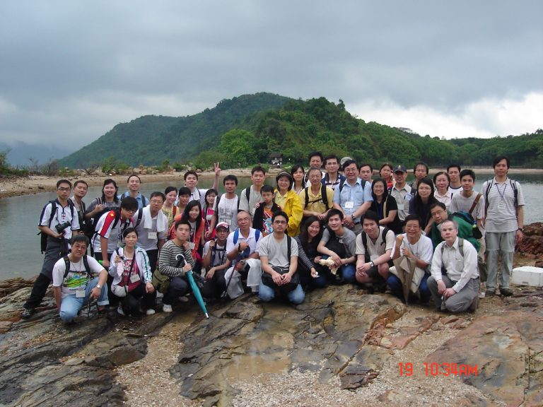 Group of people posing for photo on rocky shoreline with forested hill in background.
