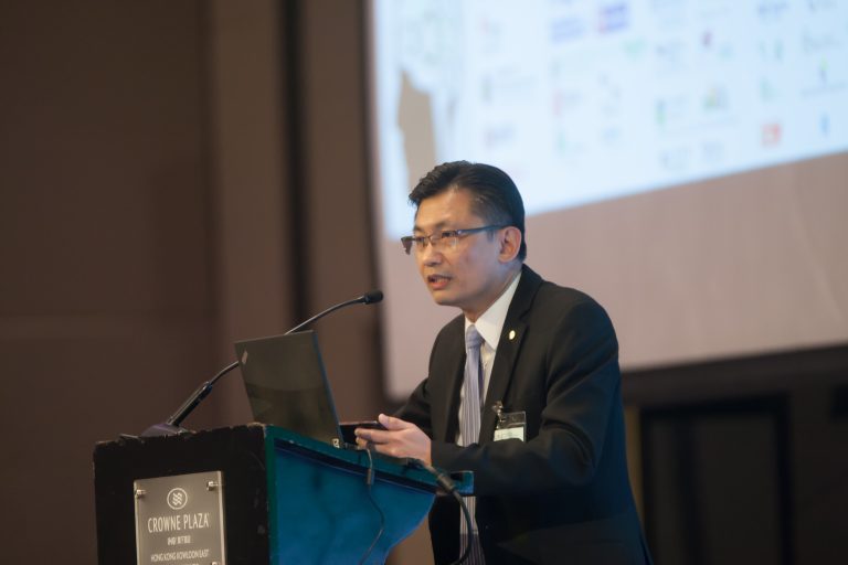Man in suit speaking at podium with laptop and presentation screen behind him.