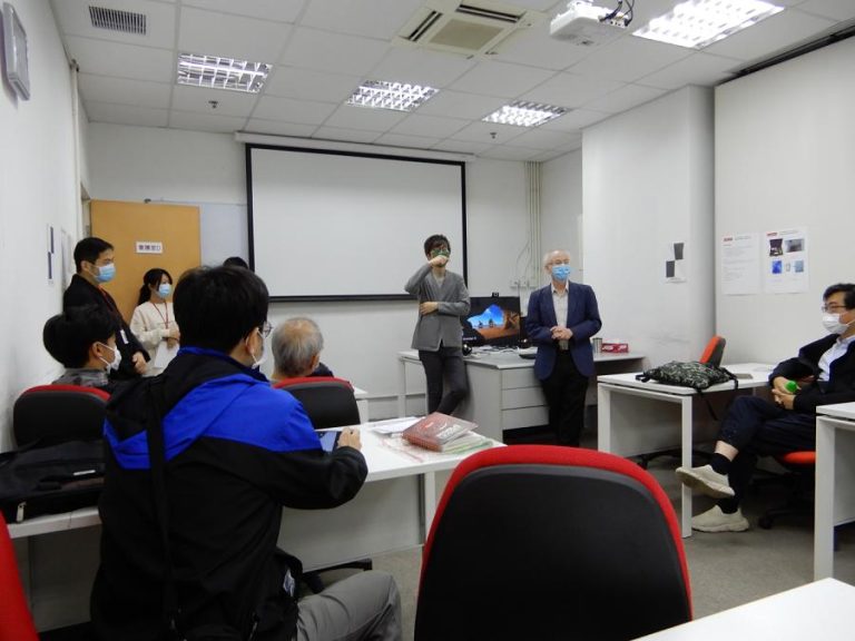People in a meeting room with masks, one standing at a presentation screen.